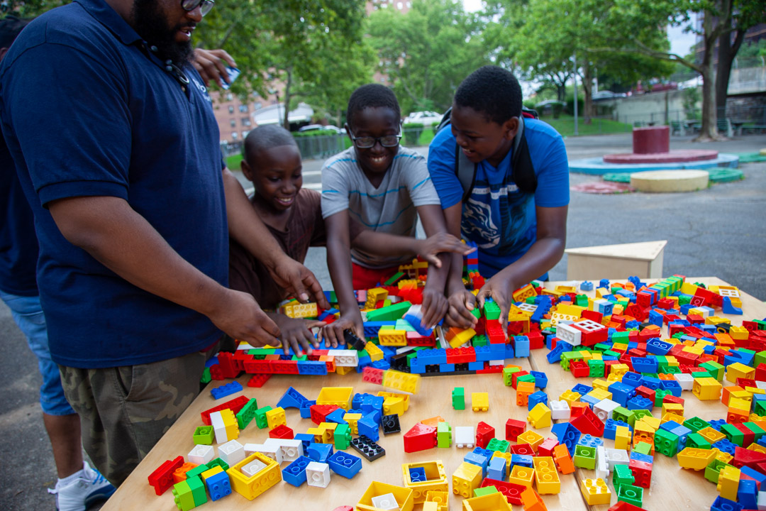 Three kids and and adult playing outdoors with Lego Duplos on a table.