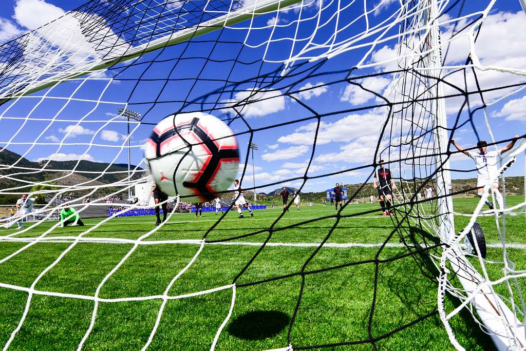 Soccer ball hitting net, with soccer players, green turf, hills, and blue sky.